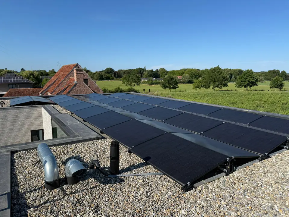 A rooftop solar panel installation on a gravel roof, with rural buildings and a green landscape in the background under a clear blue sky.