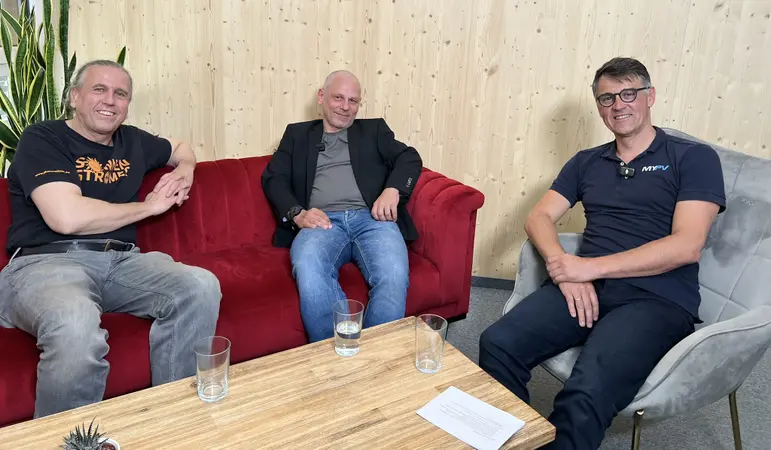 Three men on a red couch and a gray chair, smiling, with wooden wall behind and a table with glasses and papers in front