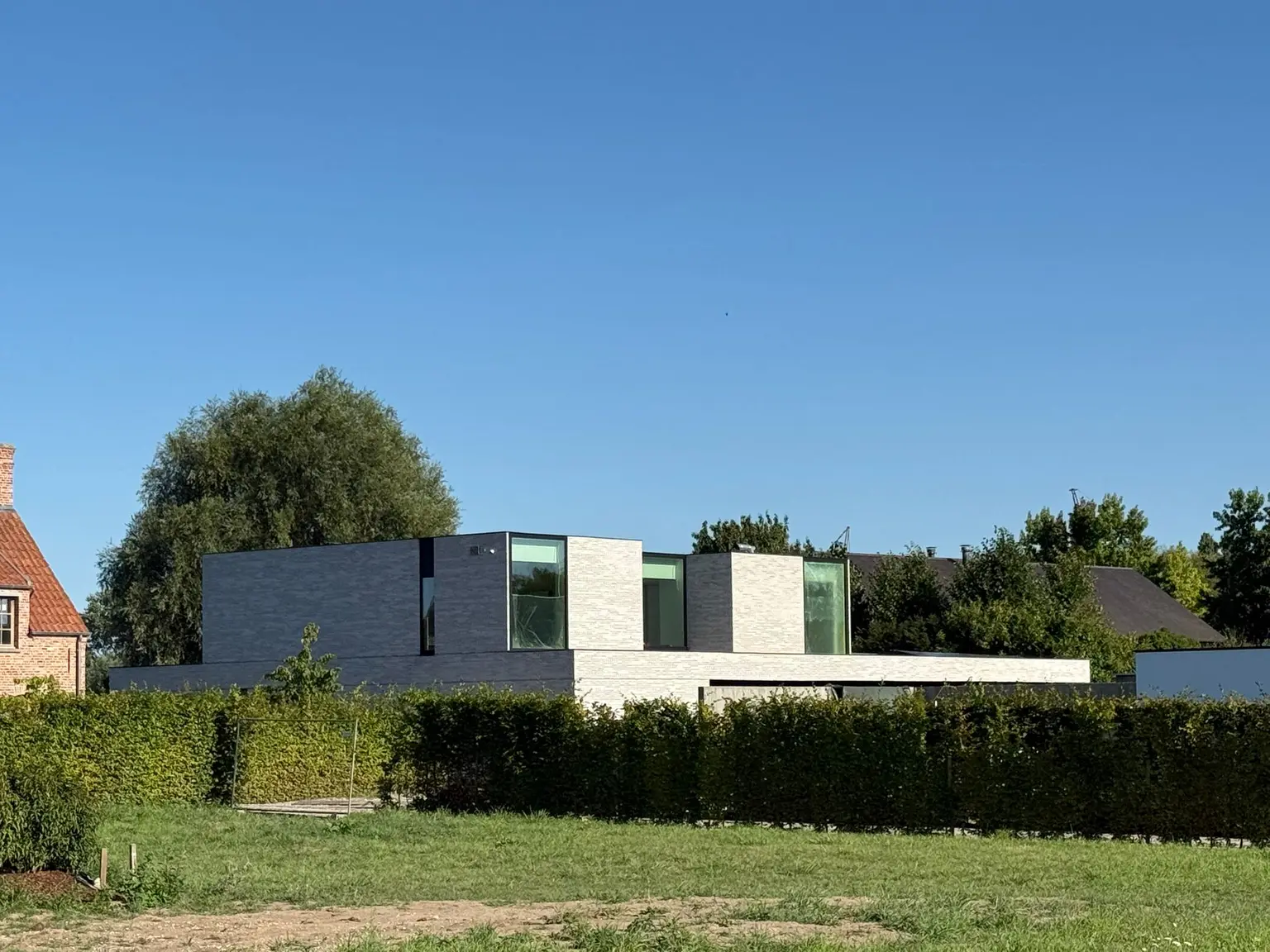 Modern flat-roofed house with large windows, surrounded by a hedge and lawn, under a clear blue sky.