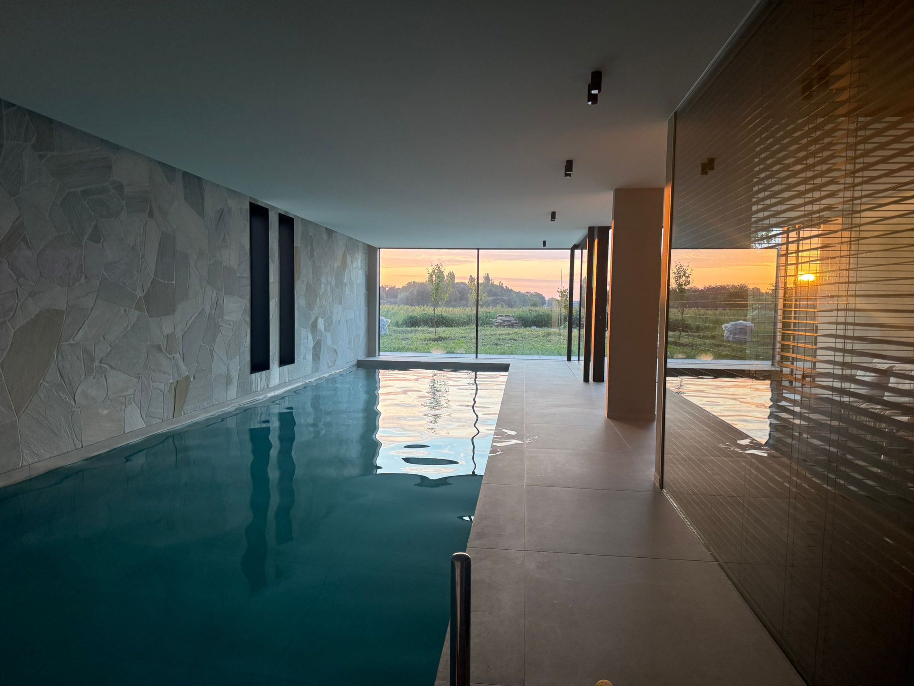 Indoor pool with large windows showing a sunset over a rural landscape. The interior features stone and wood paneling.
