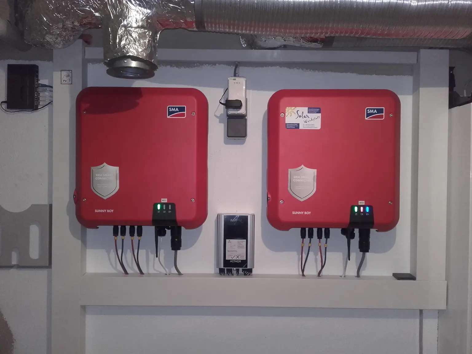 "Two red solar inverters wall-mounted, connected with cables, displaying green and red indicator lights.