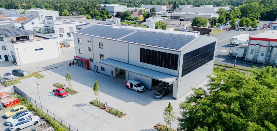 Aerial view of a modern industrial building with solar panels, surrounded by other buildings, parking spaces, and green trees.