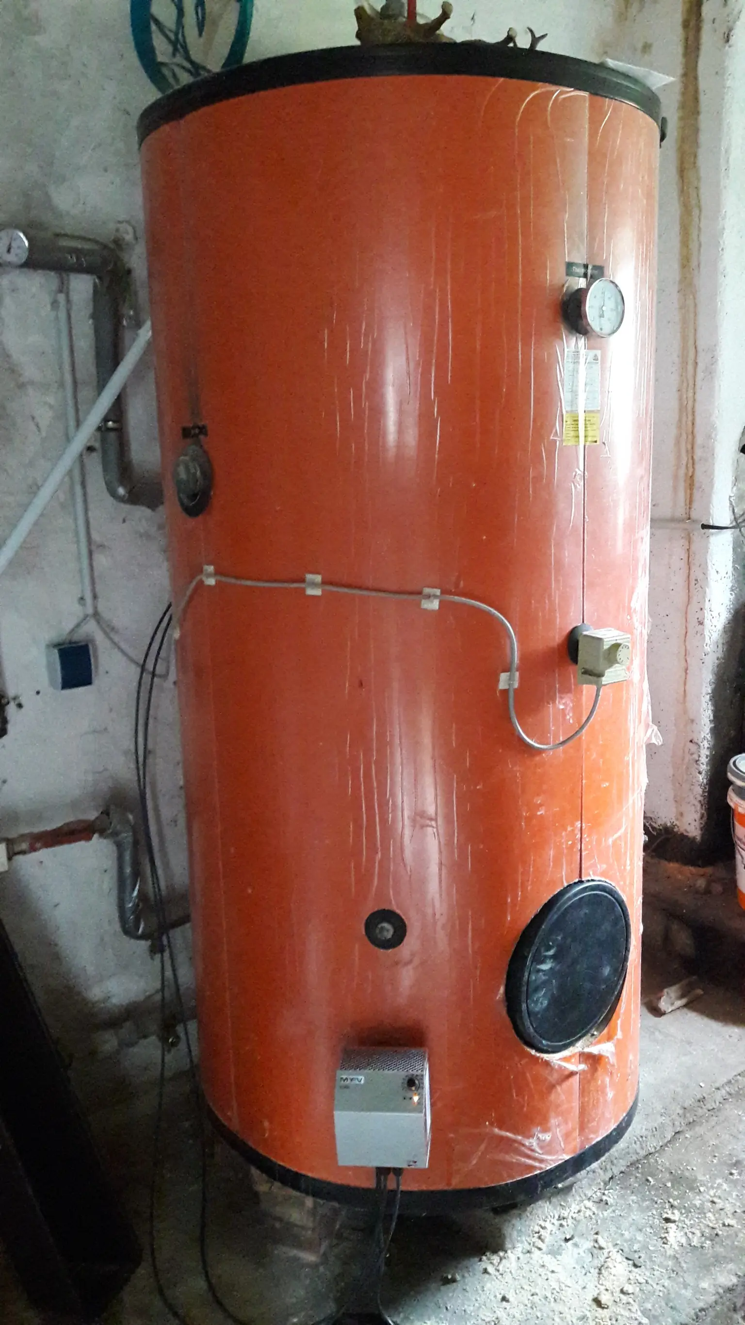 Large orange water heater tank with mounted ELWA, gauges, and pipes, placed in a utility room.