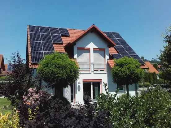 Modern house with solar panels on red-tiled roof, surrounded by lush greenery under clear blue sky.