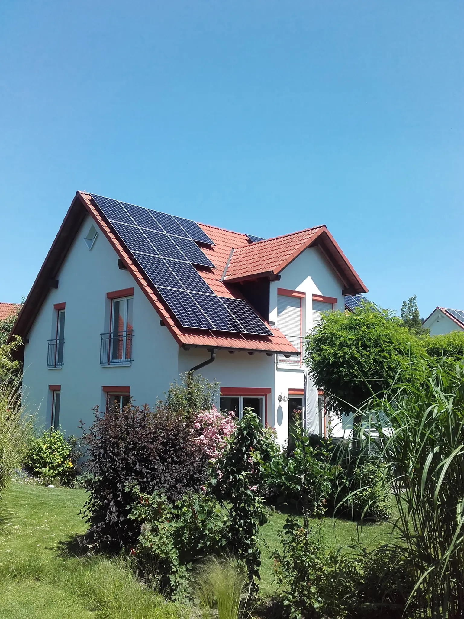 White house with red roof tiles and solar panels, surrounded by greenery and shrubs under clear blue sky.