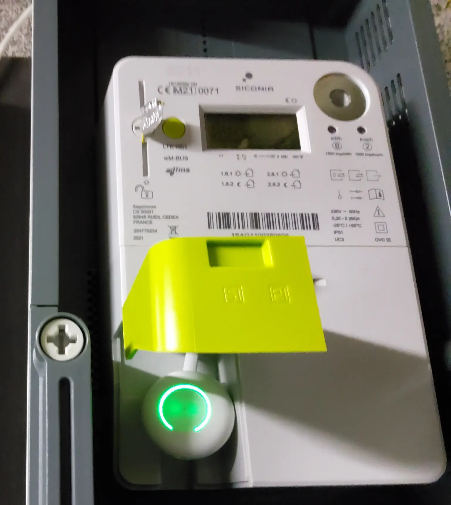 A digital electric meter with a yellow cover and a glowing green button, displaying various readings and symbols.