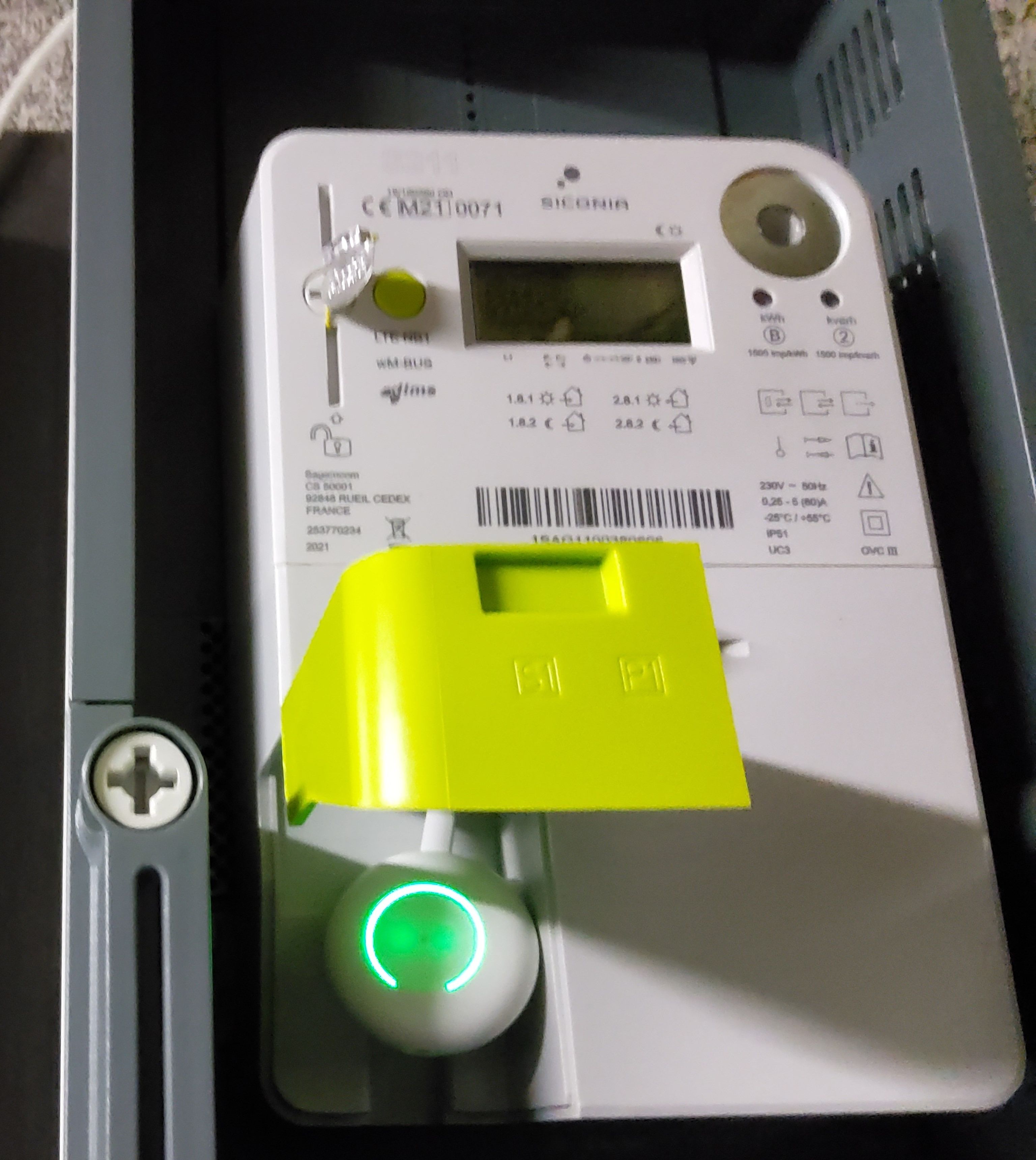 A digital electric meter with a yellow cover and a glowing green button, displaying various readings and symbols.