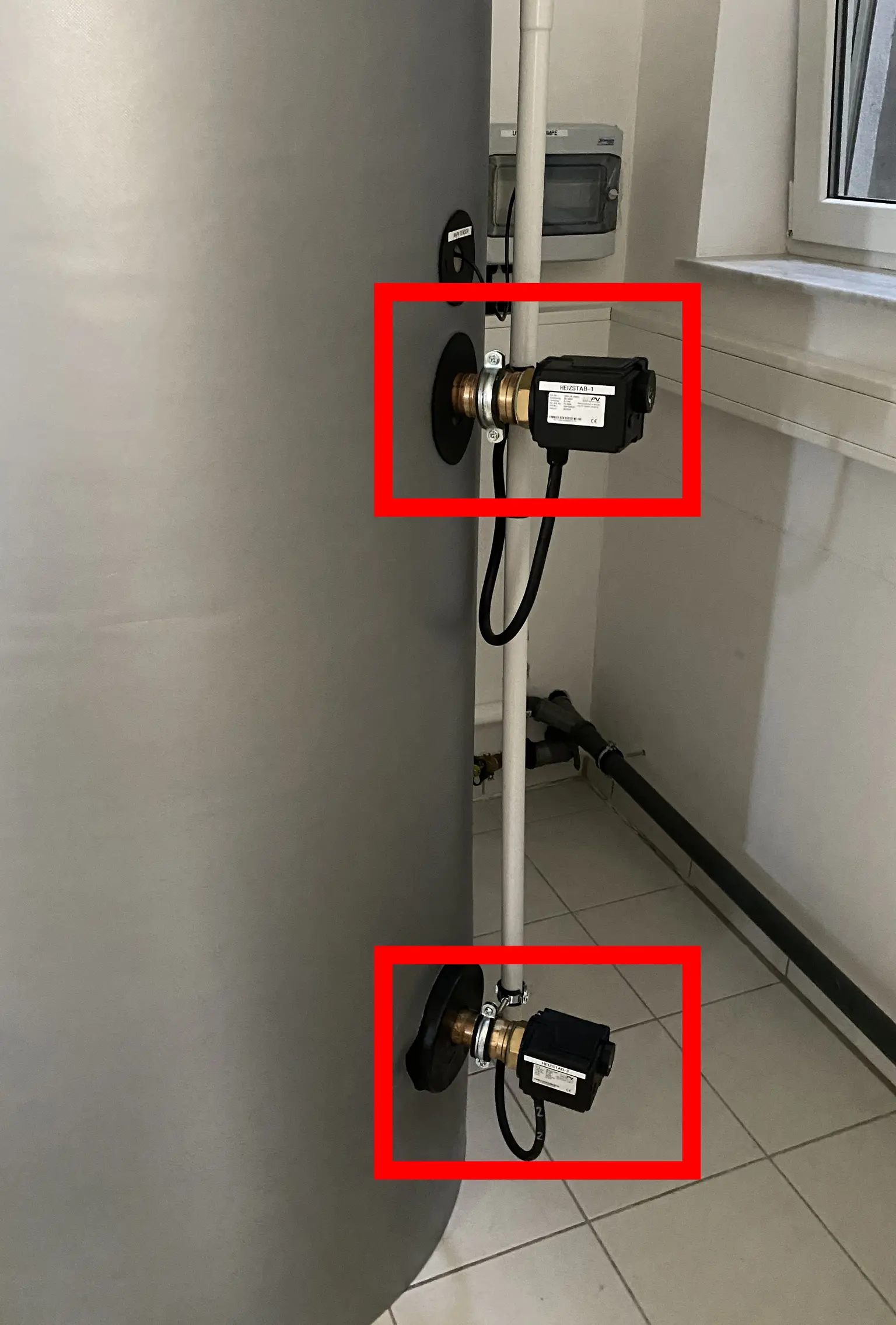 Large cylindrical tank with heating element attached to pipes, positioned vertically, with tiled floor and window in background.