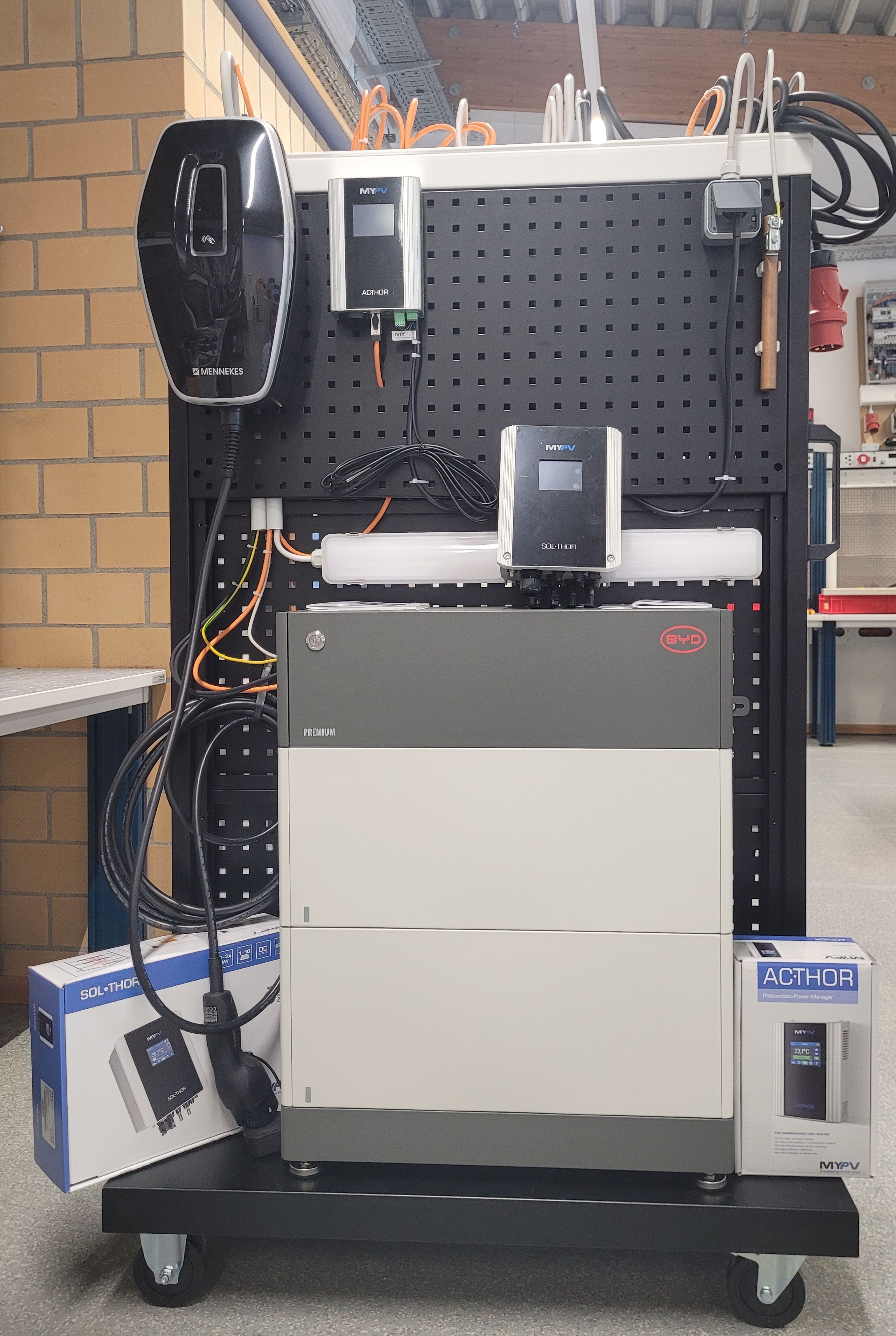 Energy management system display with a solar inverter, battery storage unit, and control panels on a mobile pegboard stand in a workshop.