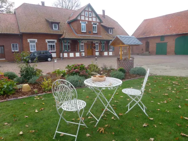 White garden chairs with a table in a green setting, some leaves on the ground, and a charming farmhouse in the background.
