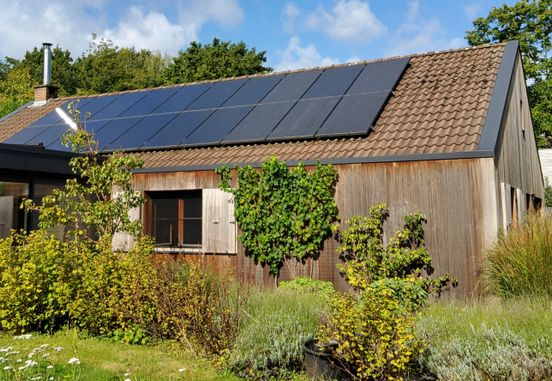 A wooden house with solar panels on the roof, surrounded by green trees and a clear blue sky.