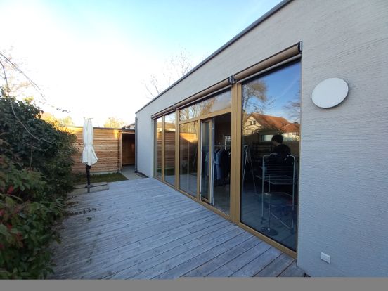 Modern house with large glass doors opening to a wooden deck, surrounded by greenery, and a white umbrella on the patio.