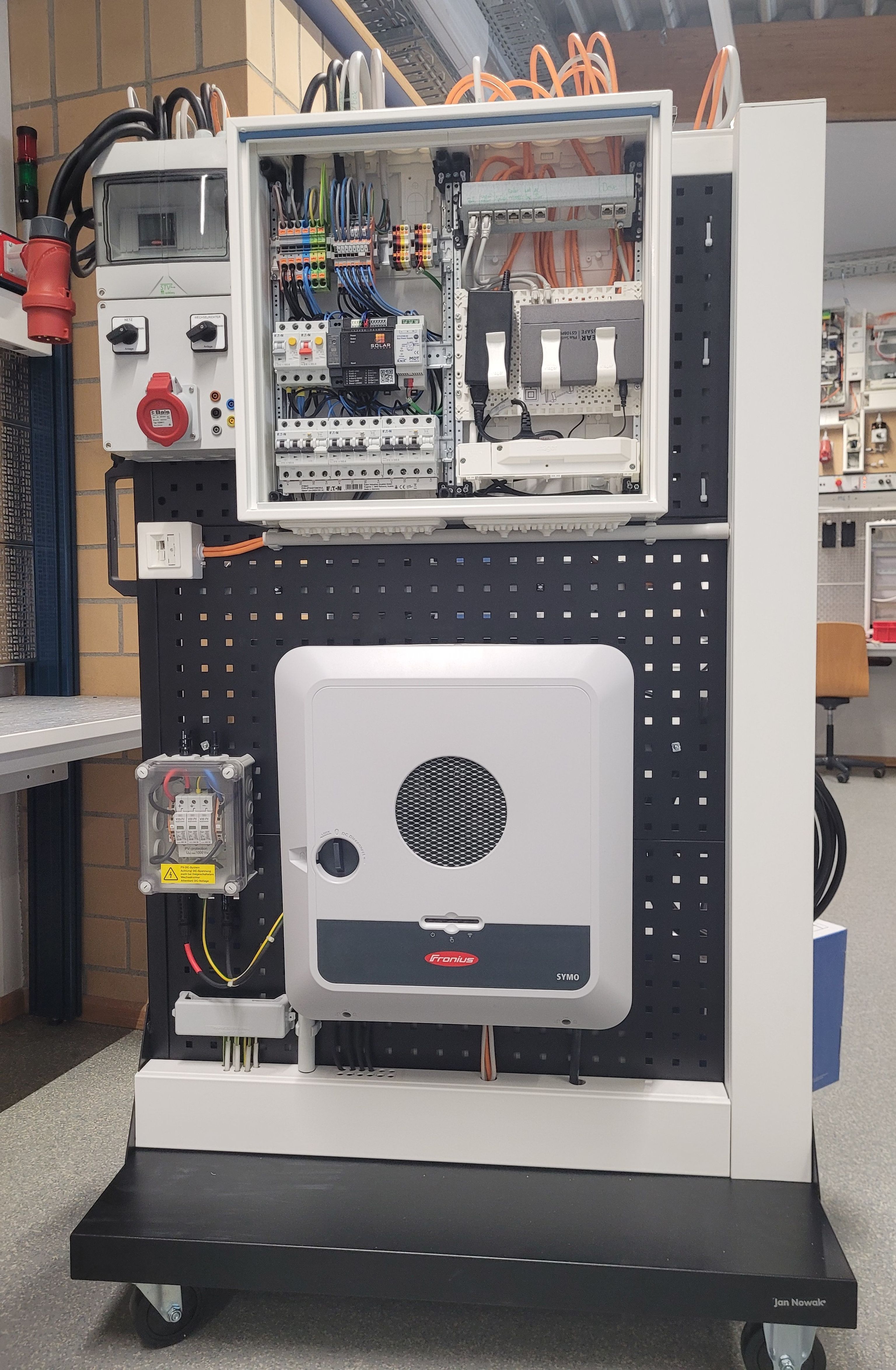 Electrical panel display with wiring, switches, and a large inverter unit on a wheeled stand in an industrial setting.