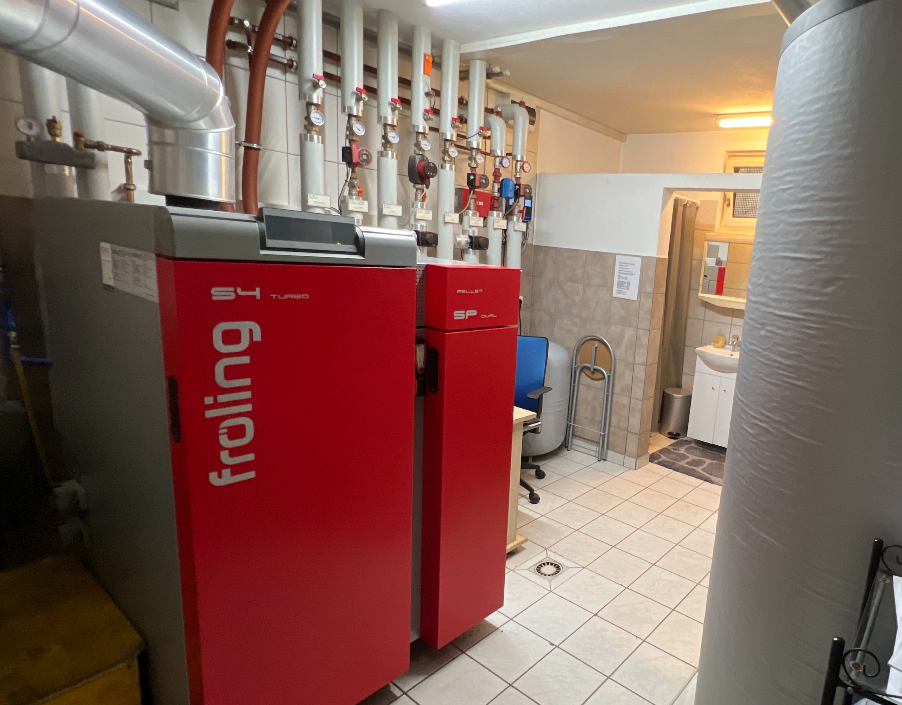 A room with red boilers, pipes, and gauges on the wall, a blue chair, and a small bathroom visible in the background.