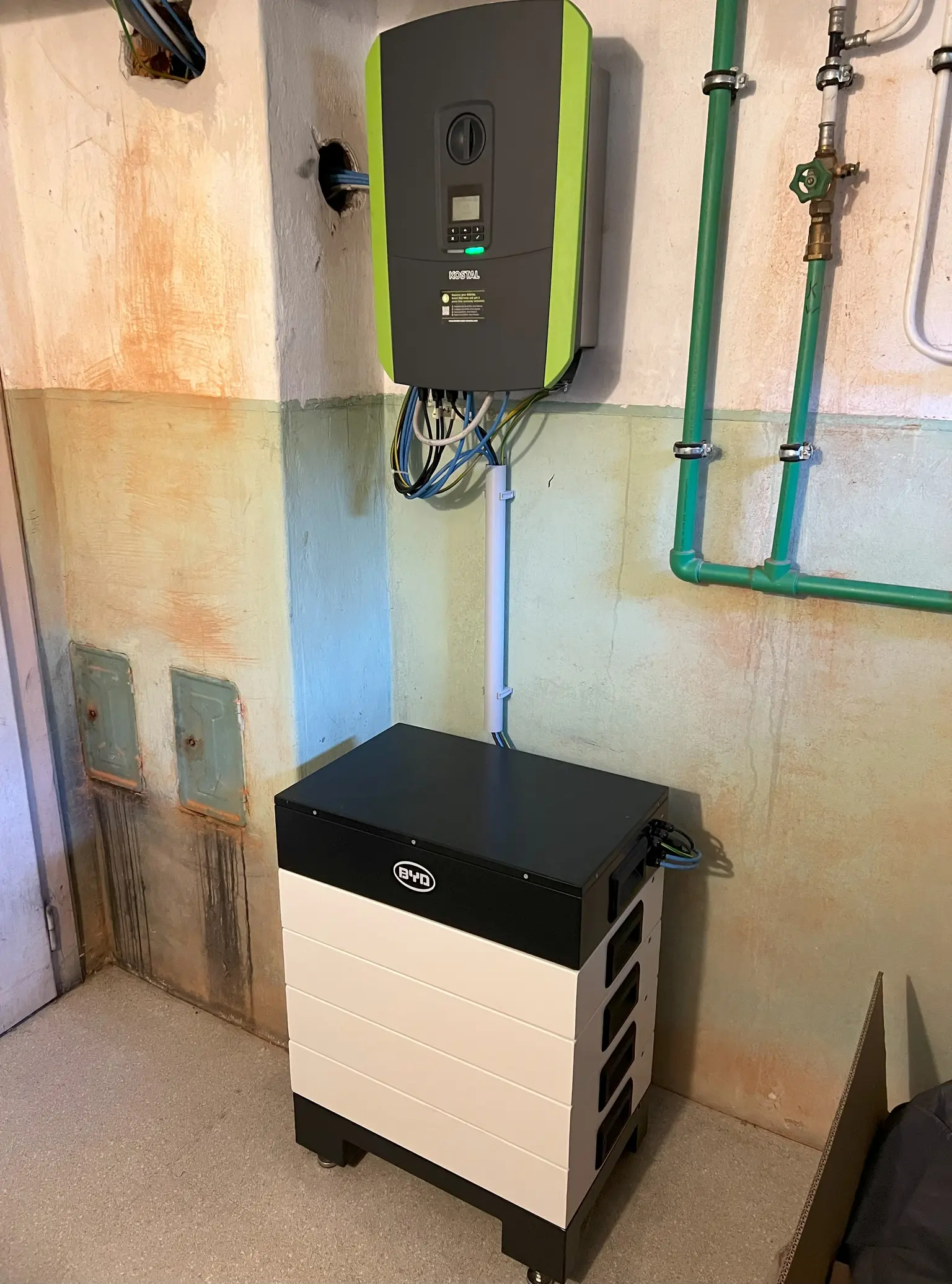 Wall-mounted Kostal inverter with many cables running out and various pipes surrounding it.