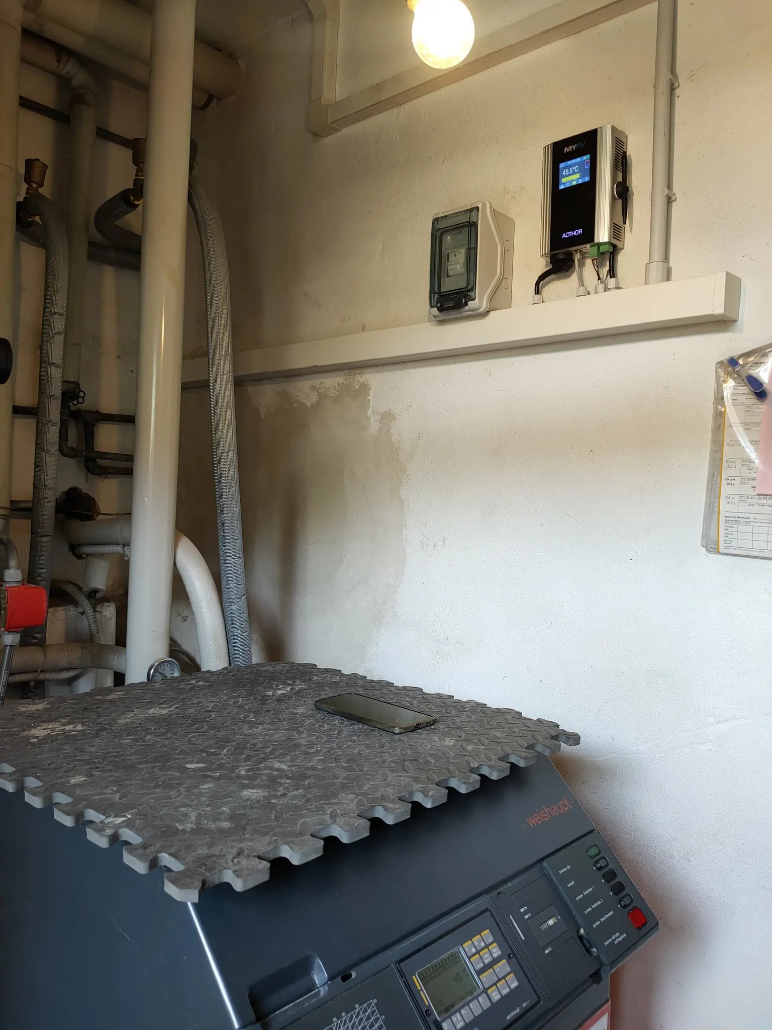 Utility room with a large machine, pipes, wall-mounted control panel, and a smartphone placed on top of the machine.