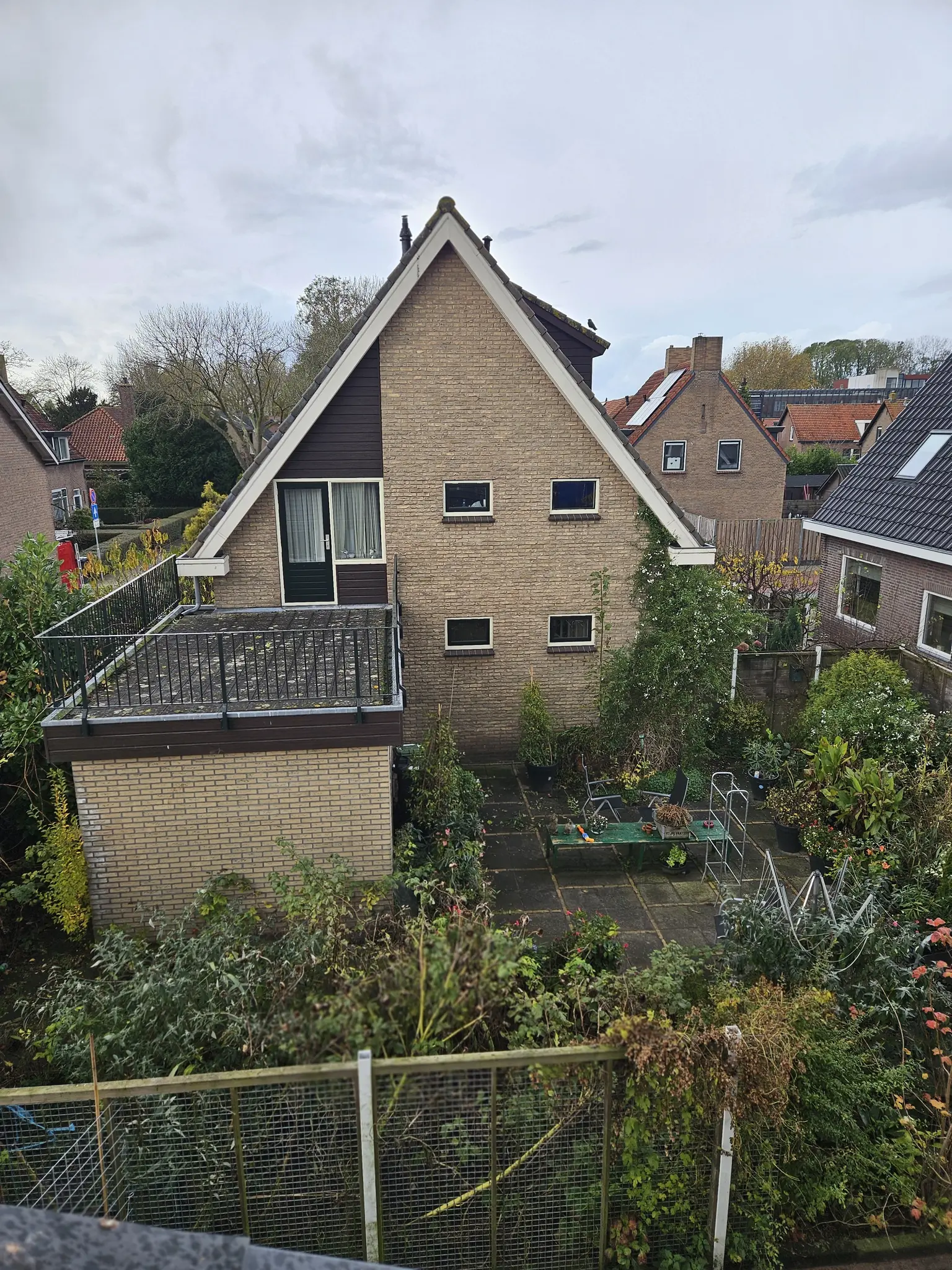 Rear view of a gabled house with a green garden and a terrace surrounded by lush vegetation.