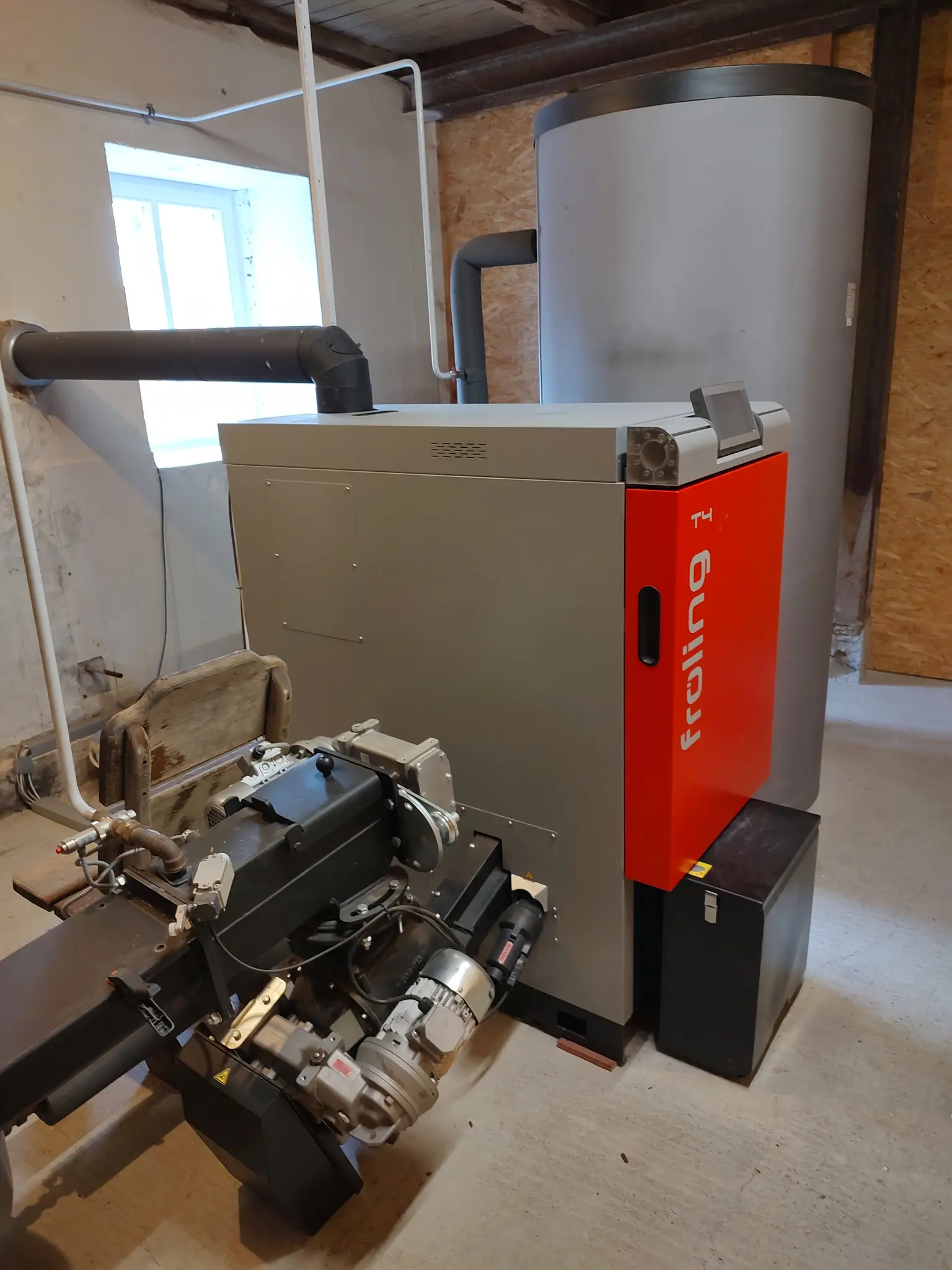Wood chip heating system in basement with connected water boiler, visible pipes and heating equipment in a utility room.