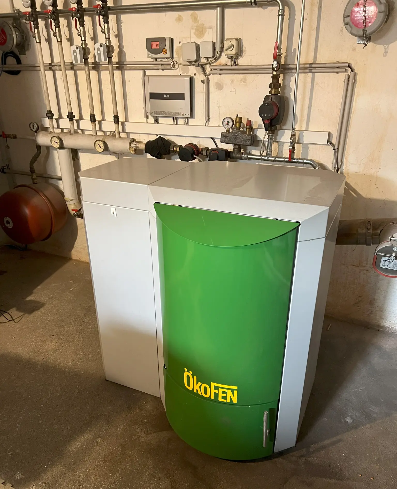 In the boiler room stands an Ökofen heating system, with several pipes and gauges visible on the wall behind it.
