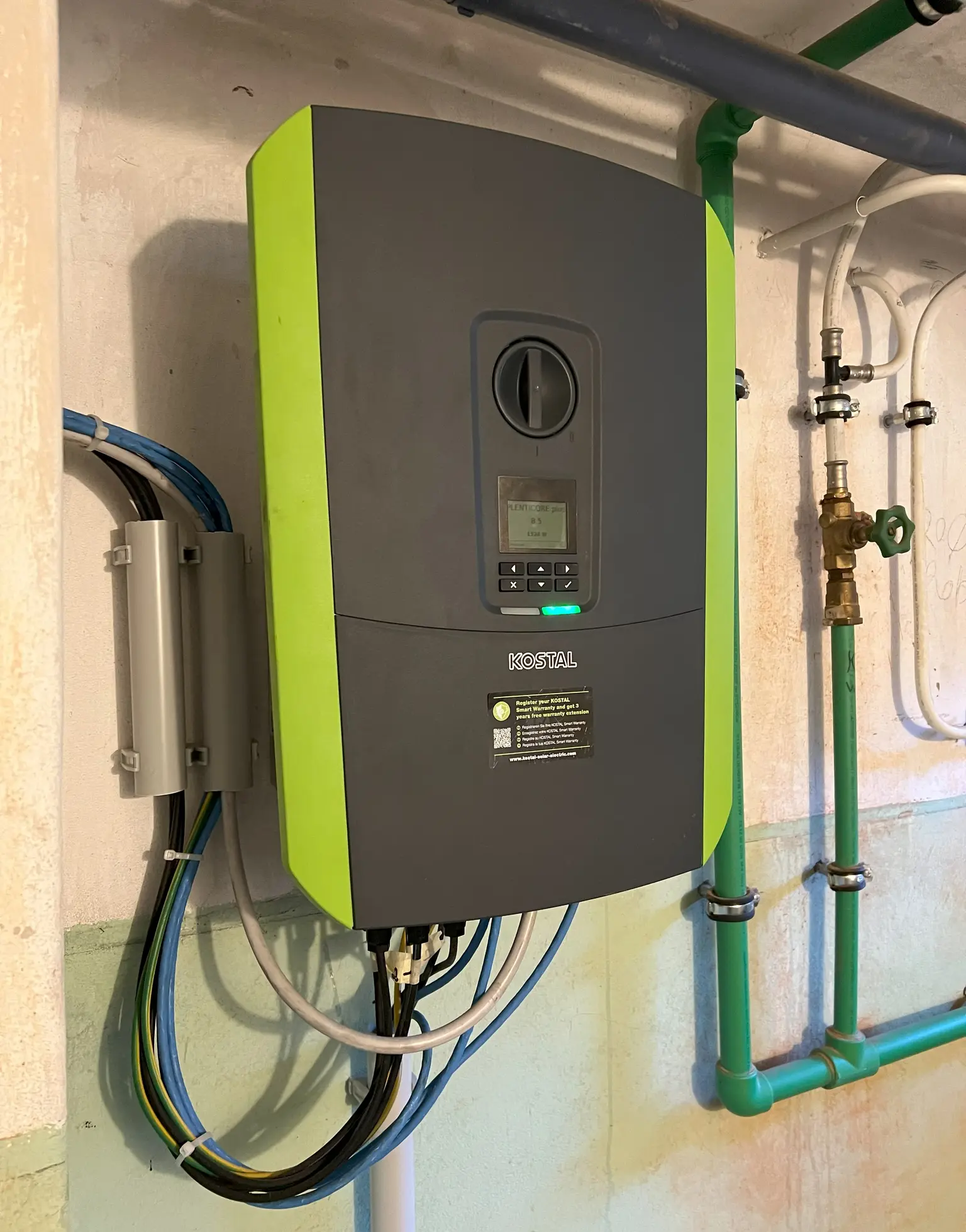 Wall-mounted Kostal inverter with many cables running out and various pipes surrounding it.