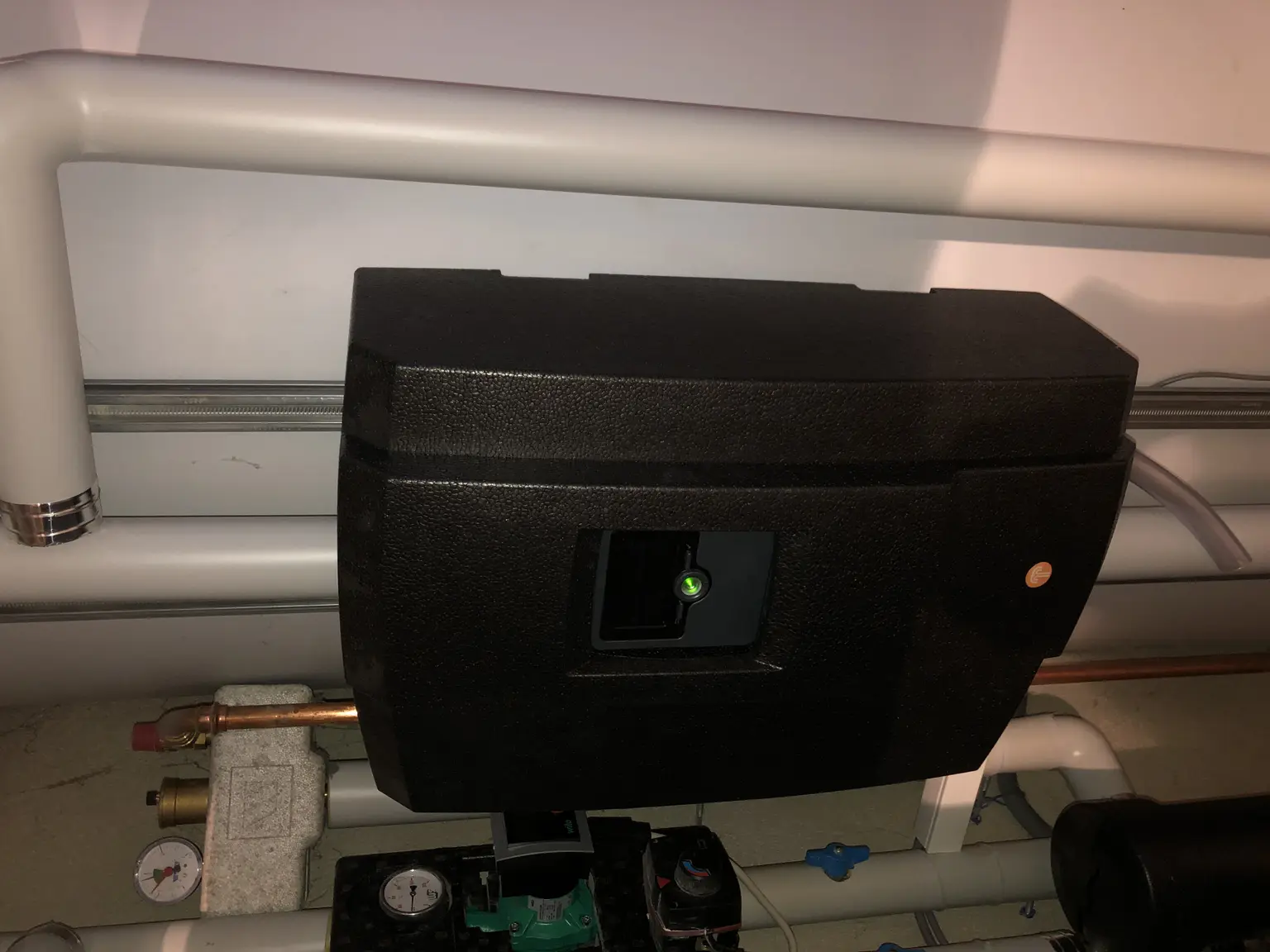 Black rectangular device wall-mounted with nearby pipes and gauges, featuring a small green indicator light.