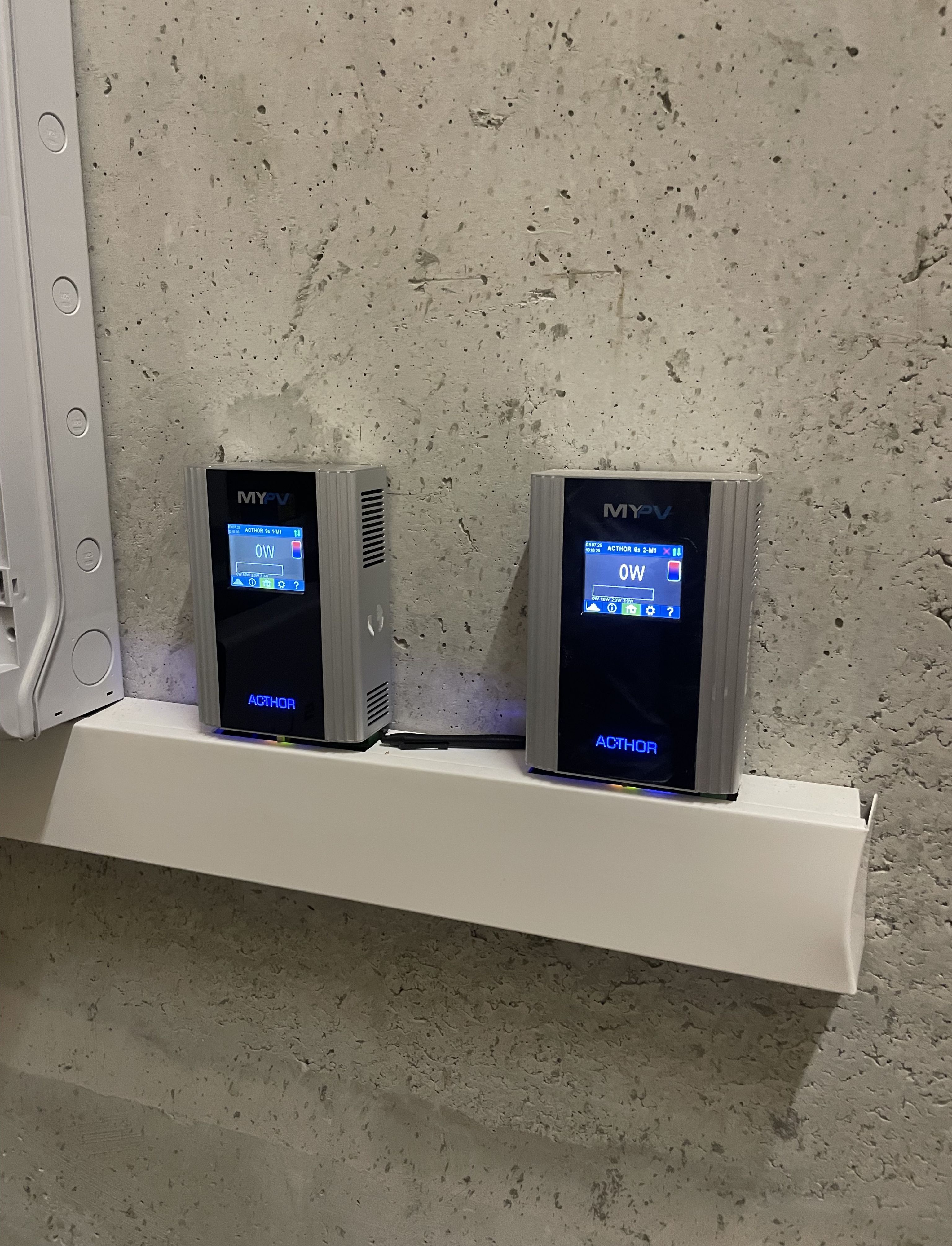 Two electronic devices with digital displays mounted on a concrete wall, showing "0W" and labeled "MY-PV" and "AC•THOR."