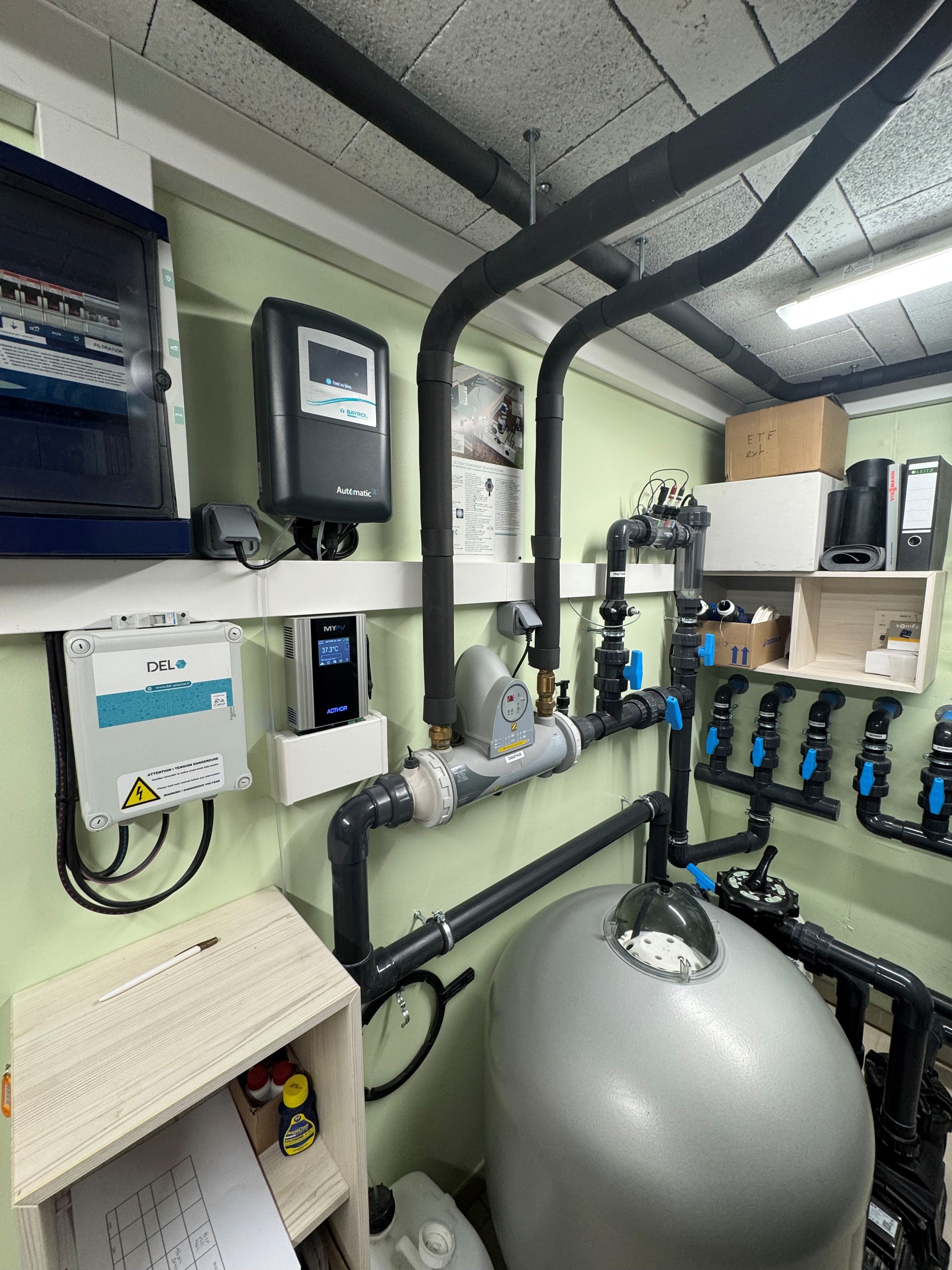 Utility room with complex plumbing, control panels, large water tank, and pipes and electrical boxes on the walls.