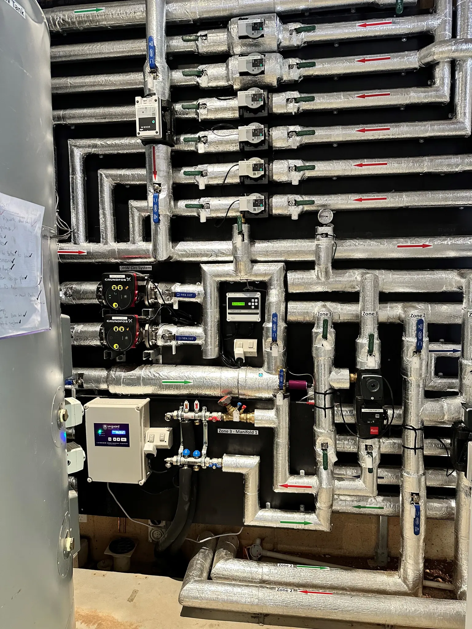 Complex network of insulated pipes with gauges, meters, and control boxes on a wall, forming a technical setup.