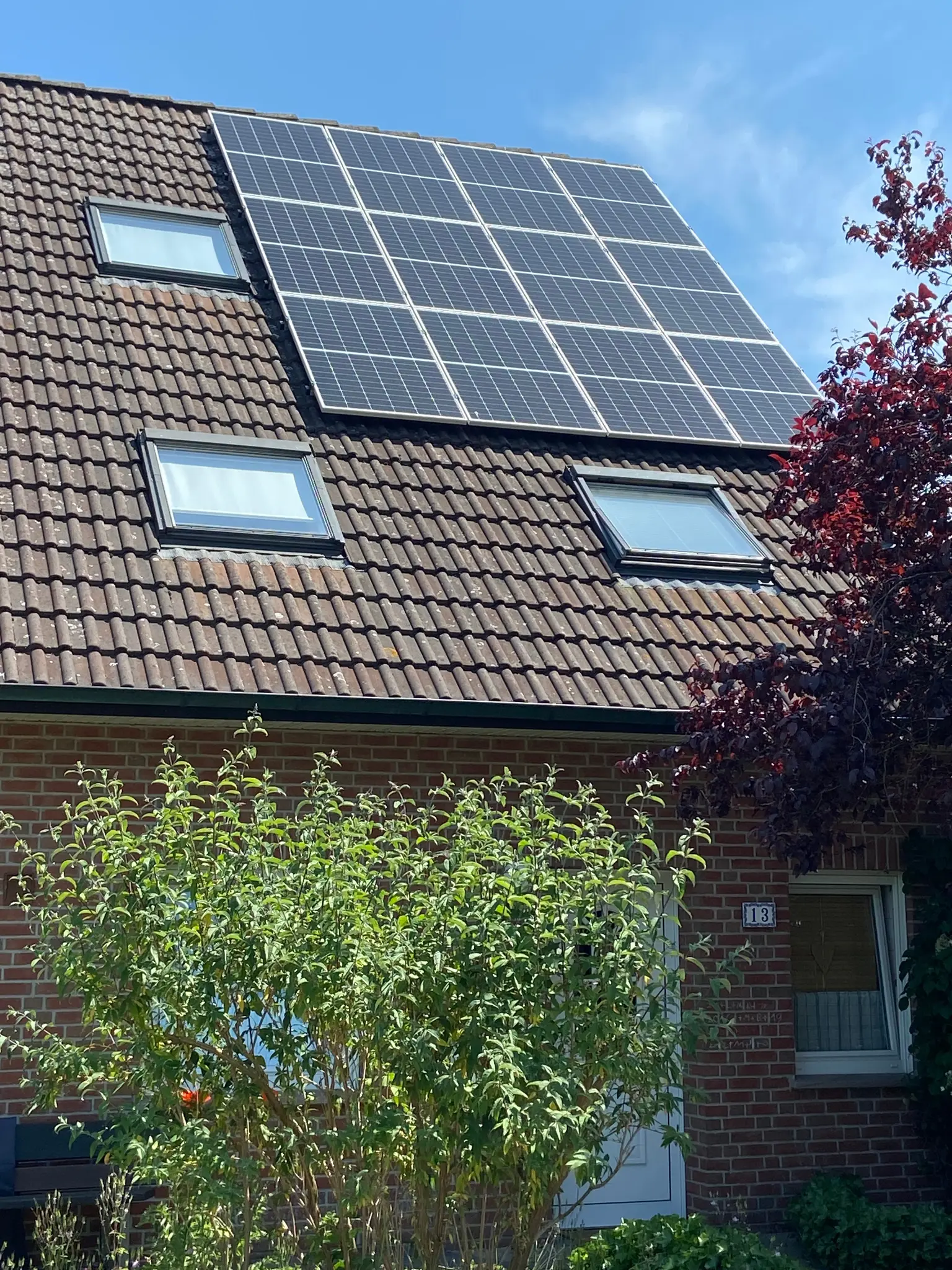 House with a brick facade features photovoltaic panels on the roof with sunlight and greenery in the foreground
