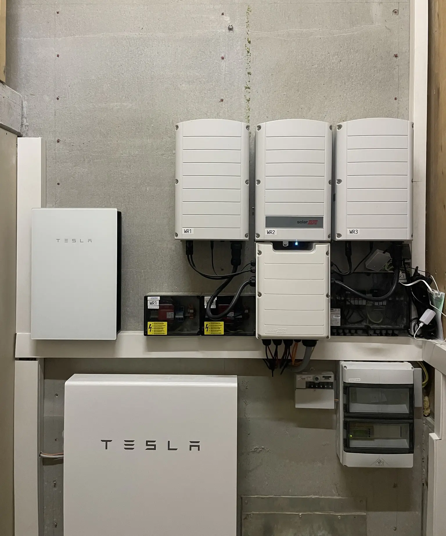 Wall-mounted Tesla Powerwall and inverters in a utility room, with cables and electrical panels visible.