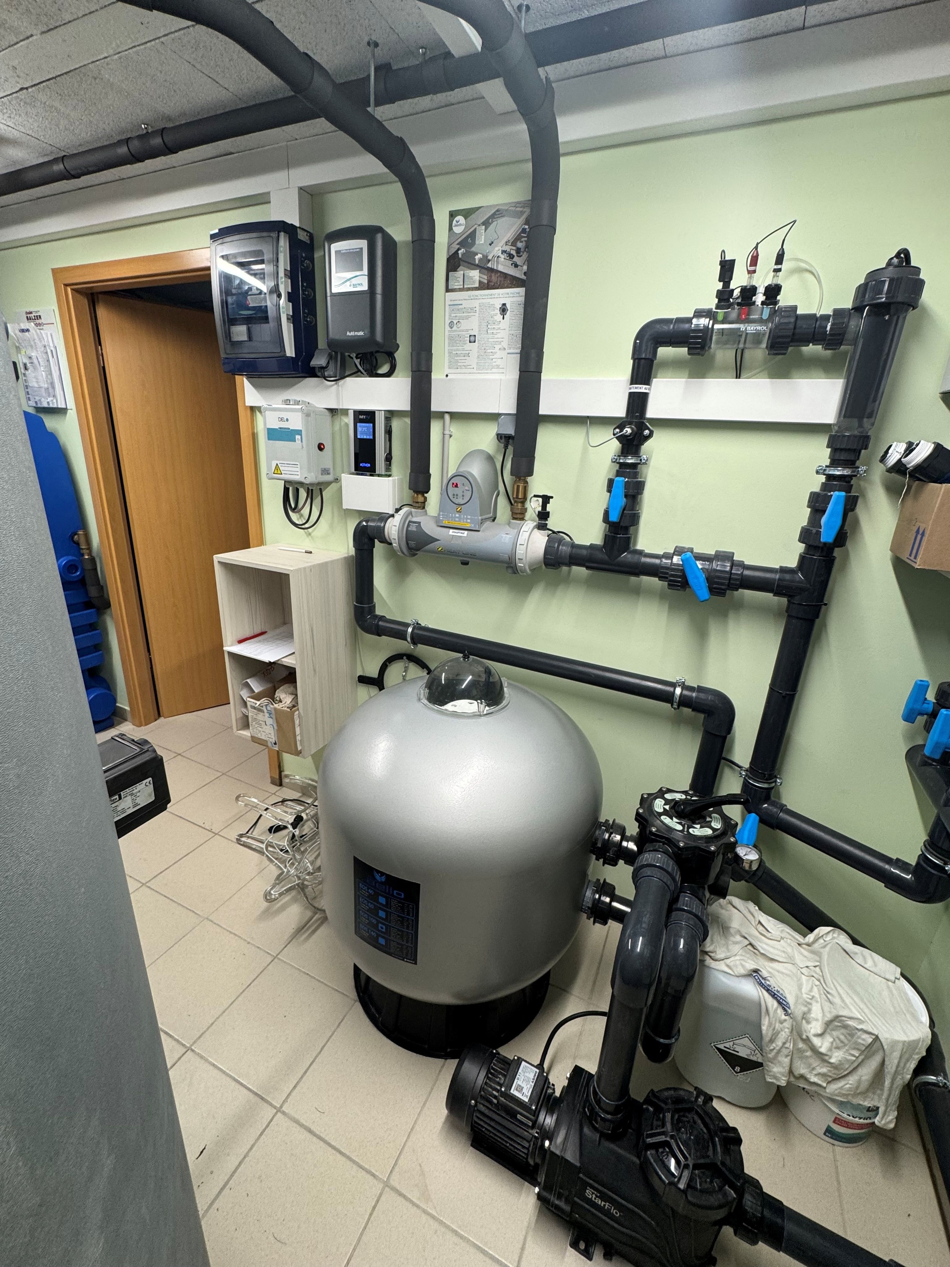 Utility room with plumbing pipes, large silver water tank, control panels, and pump system on a tiled floor.