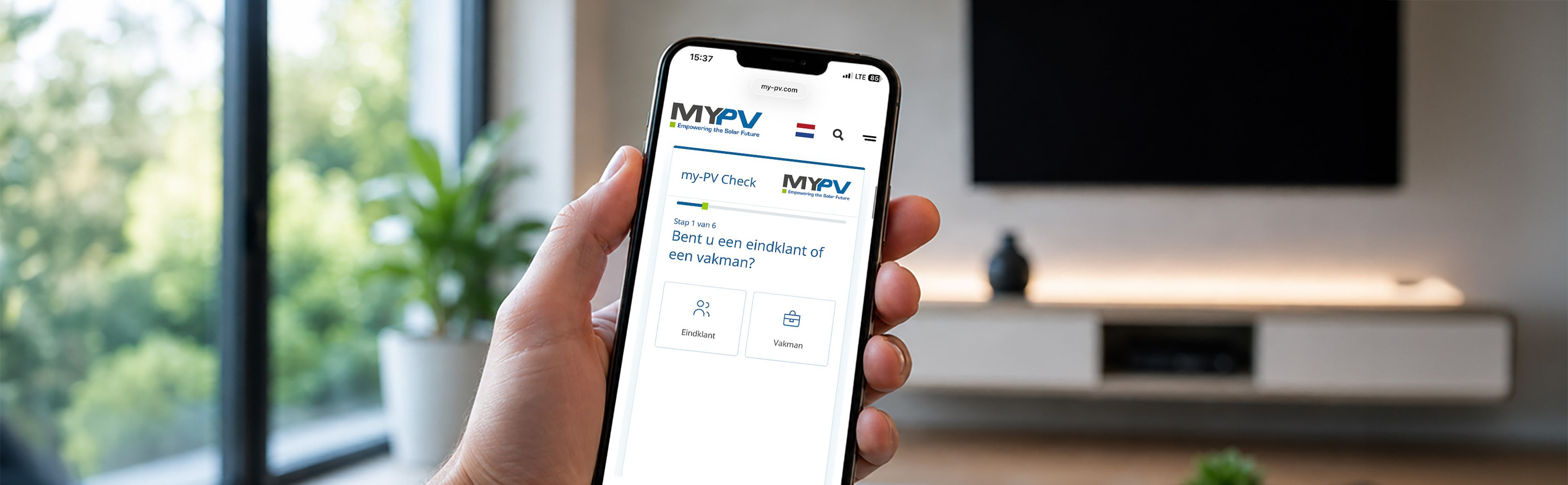 Hand holding a smartphone displaying the MY-PV solar website page, with a modern living room and greenery visible in the background.