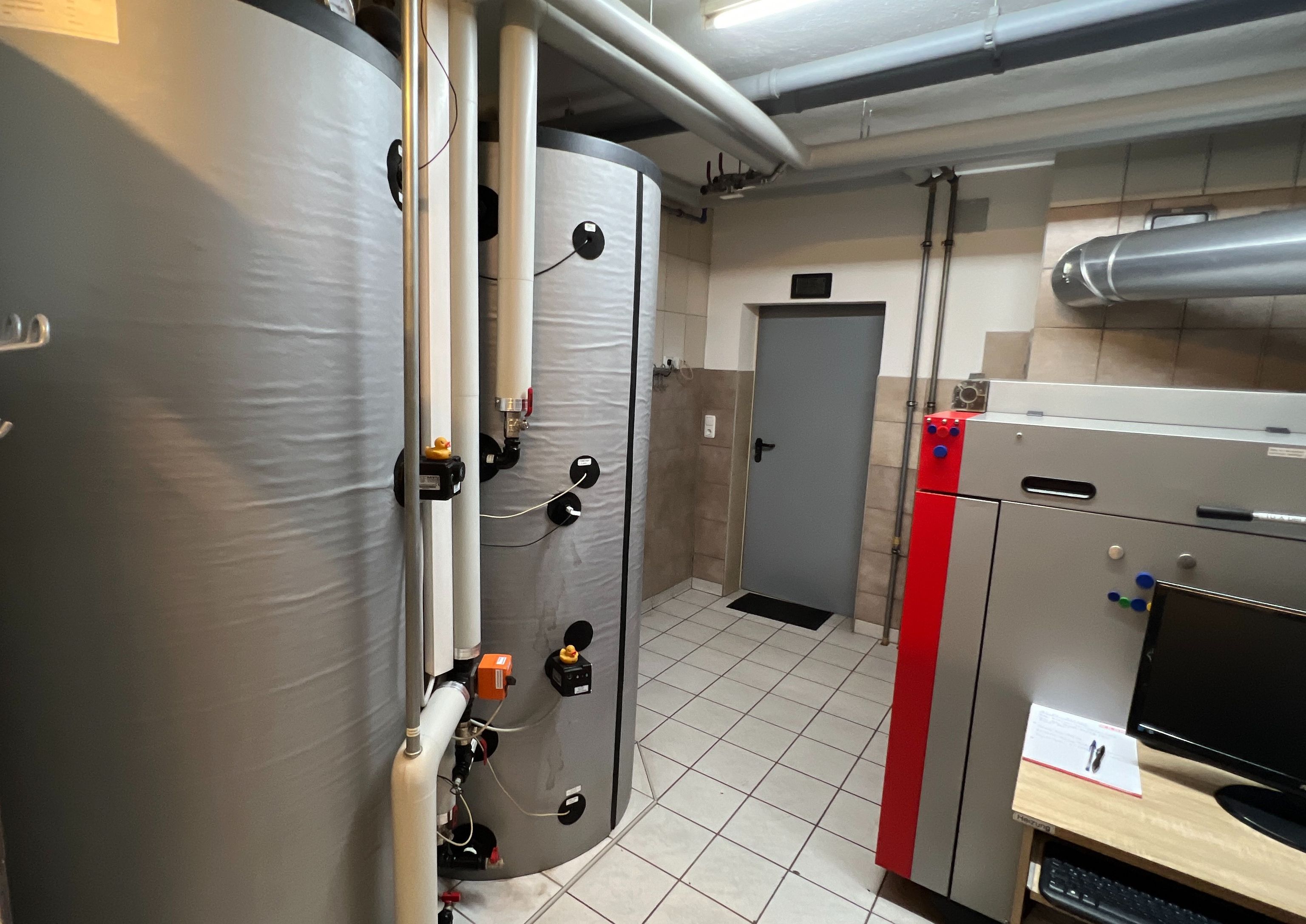 A utility room with two large insulated water tanks, pipes, and a red and gray boiler on a tiled floor.