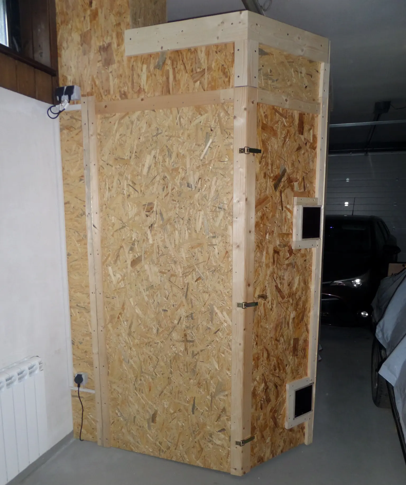Wooden enclosure with particle board panels, two small square openings, and hinges, in a garage near a parked car.