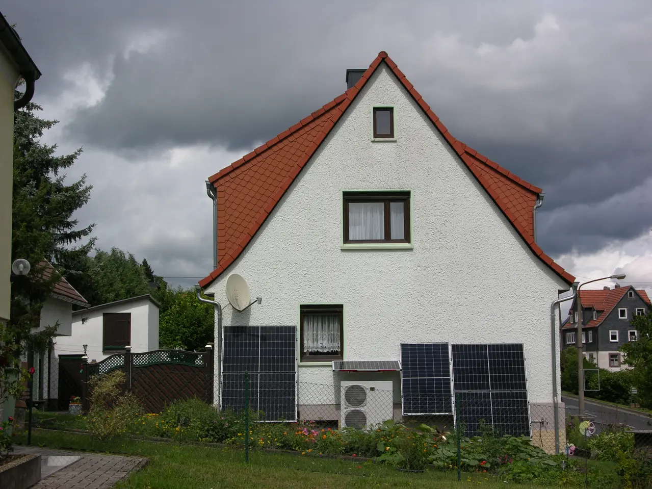 House with pointed roof, white facade, brown-framed windows, and PV panels; cloudy sky and other houses in background.