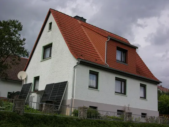 House with pointed roof, white facade, brown-framed windows, and PV panels; cloudy sky and other houses in background.