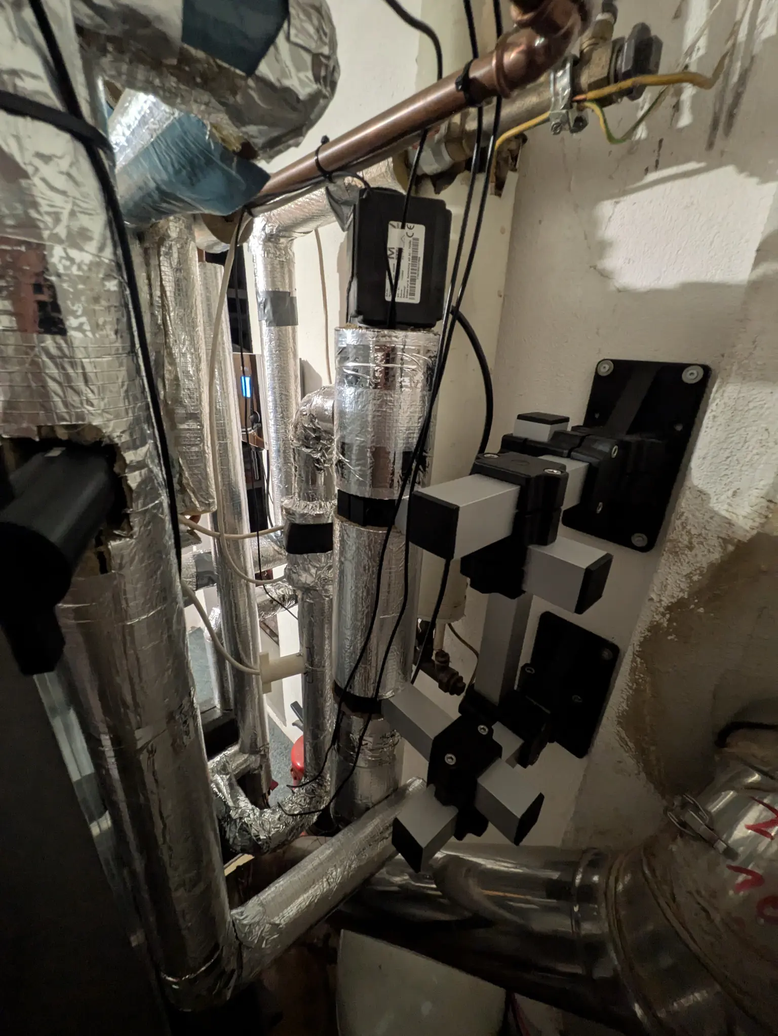 Complex network of insulated pipes and ducts in a small utility room, with cables and connectors attached to the walls.