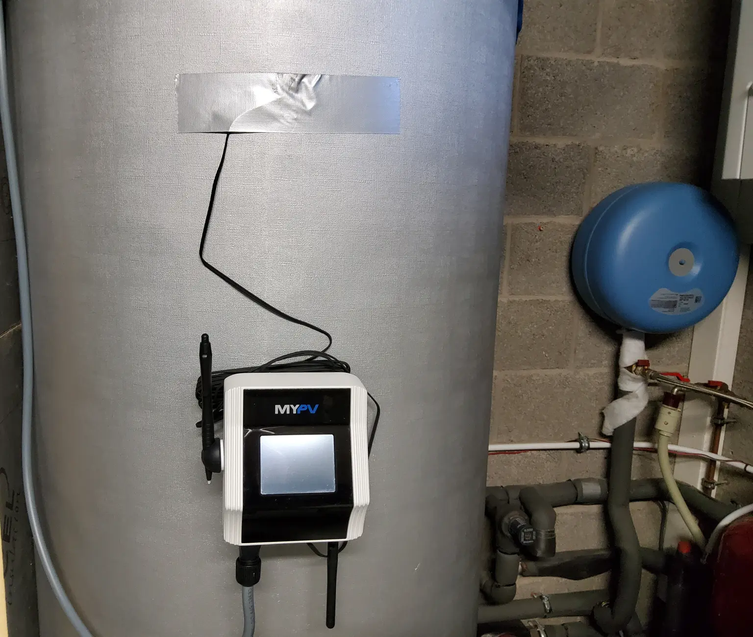 Large cylindrical water heater with an AC ELWA 2 attached, labeled, in utility room with pipes and blue pressure tank