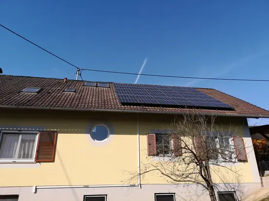 House with yellow facade and brown roof featuring solar panels, several windows, and a leafless tree under clear blue sky.