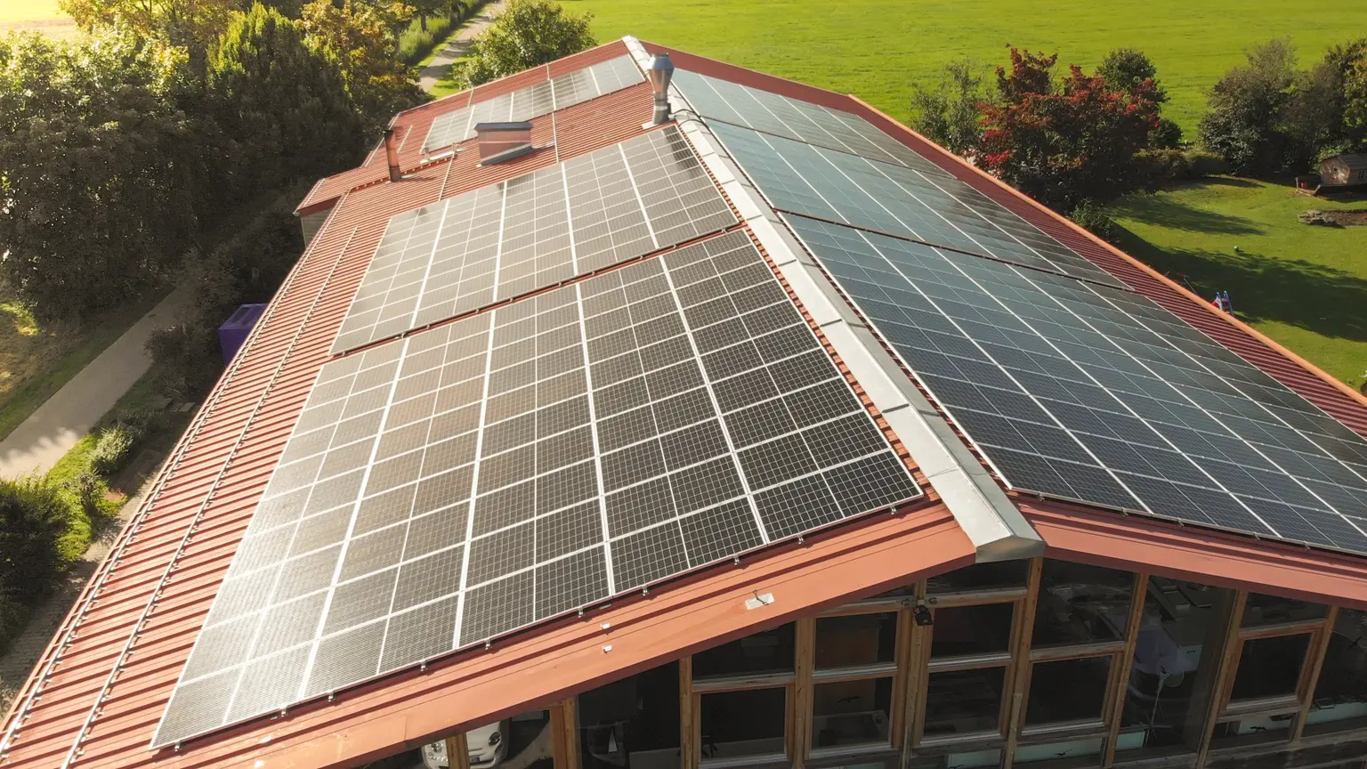 Aerial view of a building with a red roof covered in solar panels, surrounded by green fields and trees.