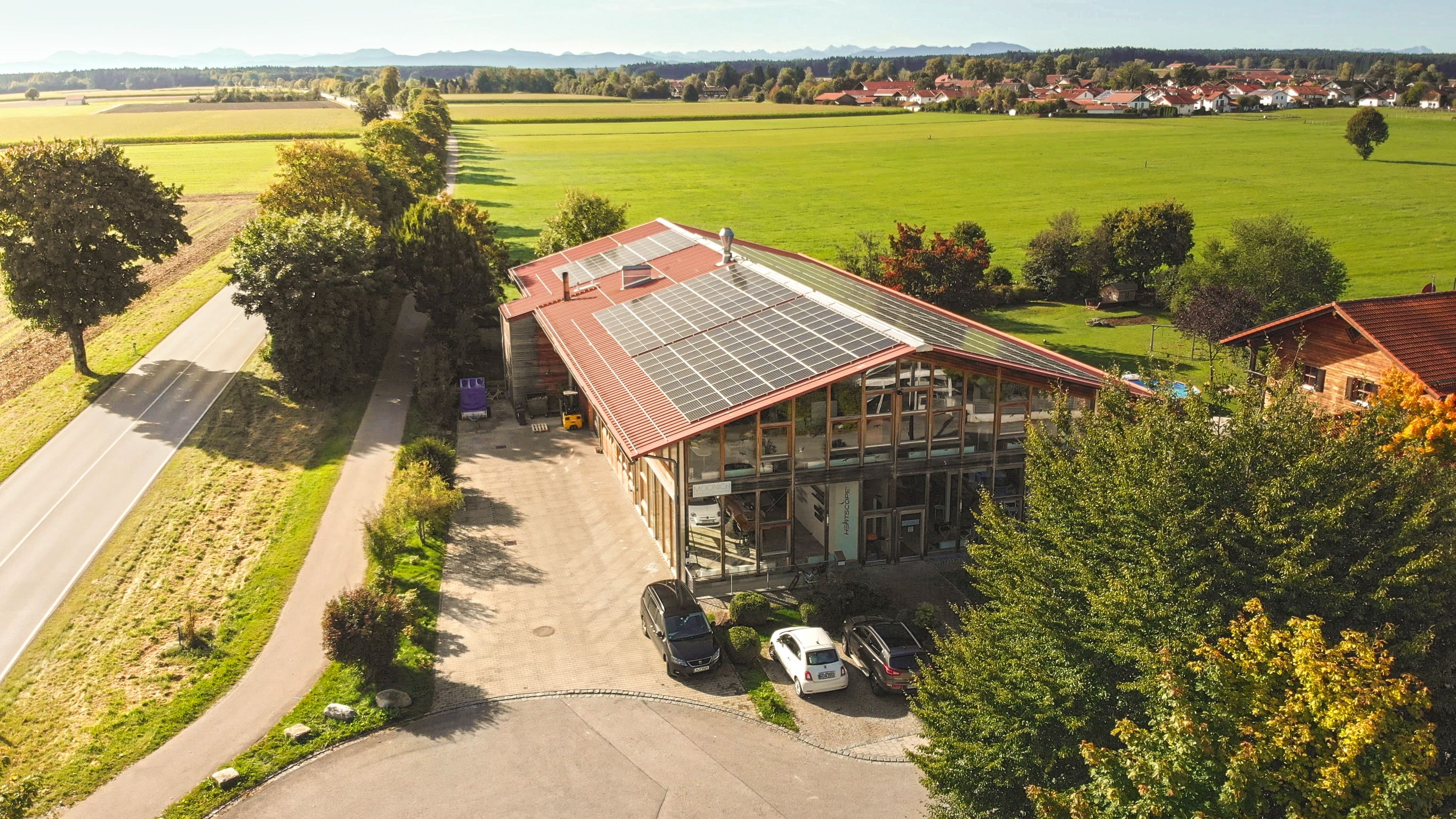 Aerial view of a building with solar panels on the roof, surrounded by green fields, a road, and trees under a clear blue sky.