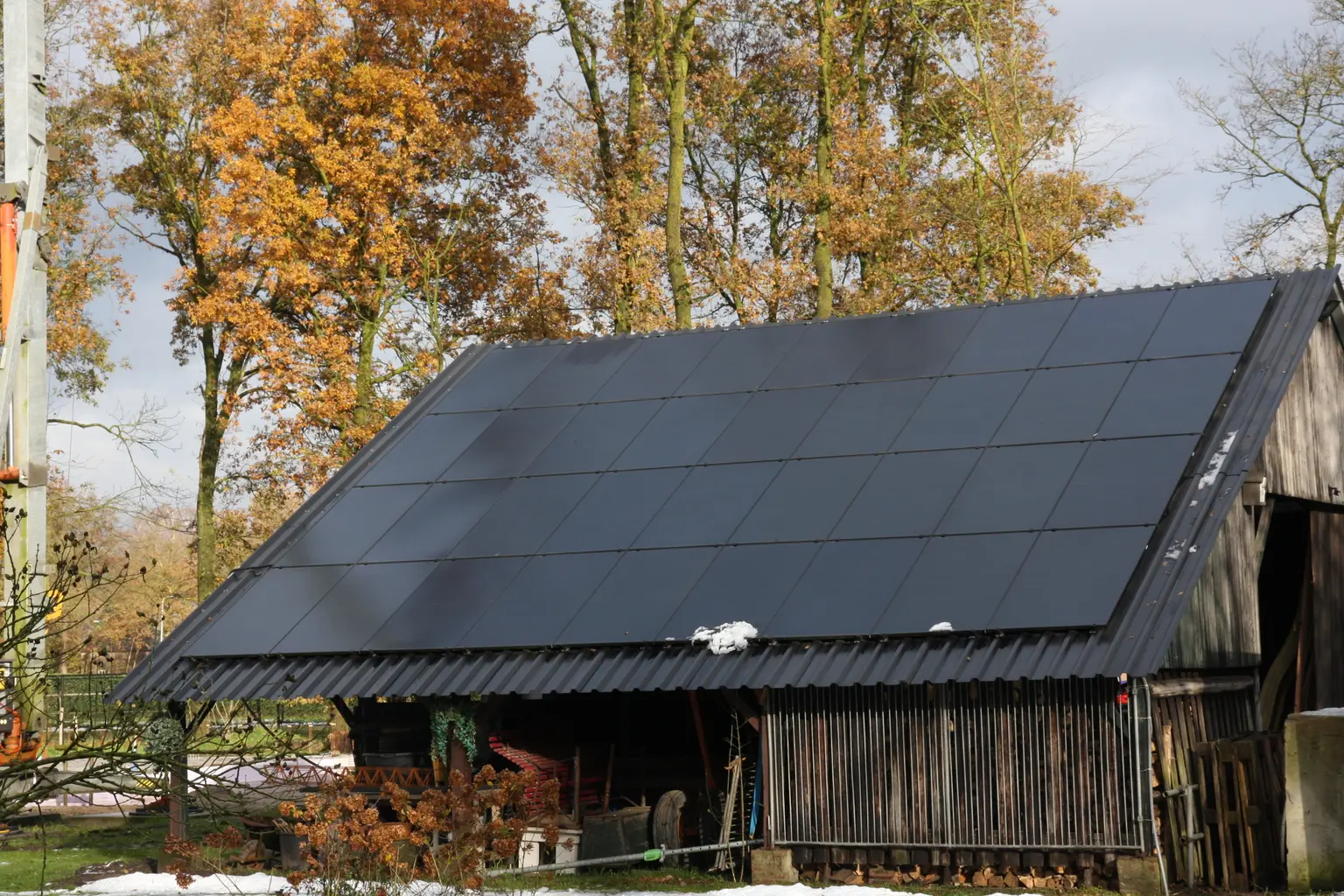 Roof of a large shed with PV panels and some snow, with autumn-orange trees in the background.