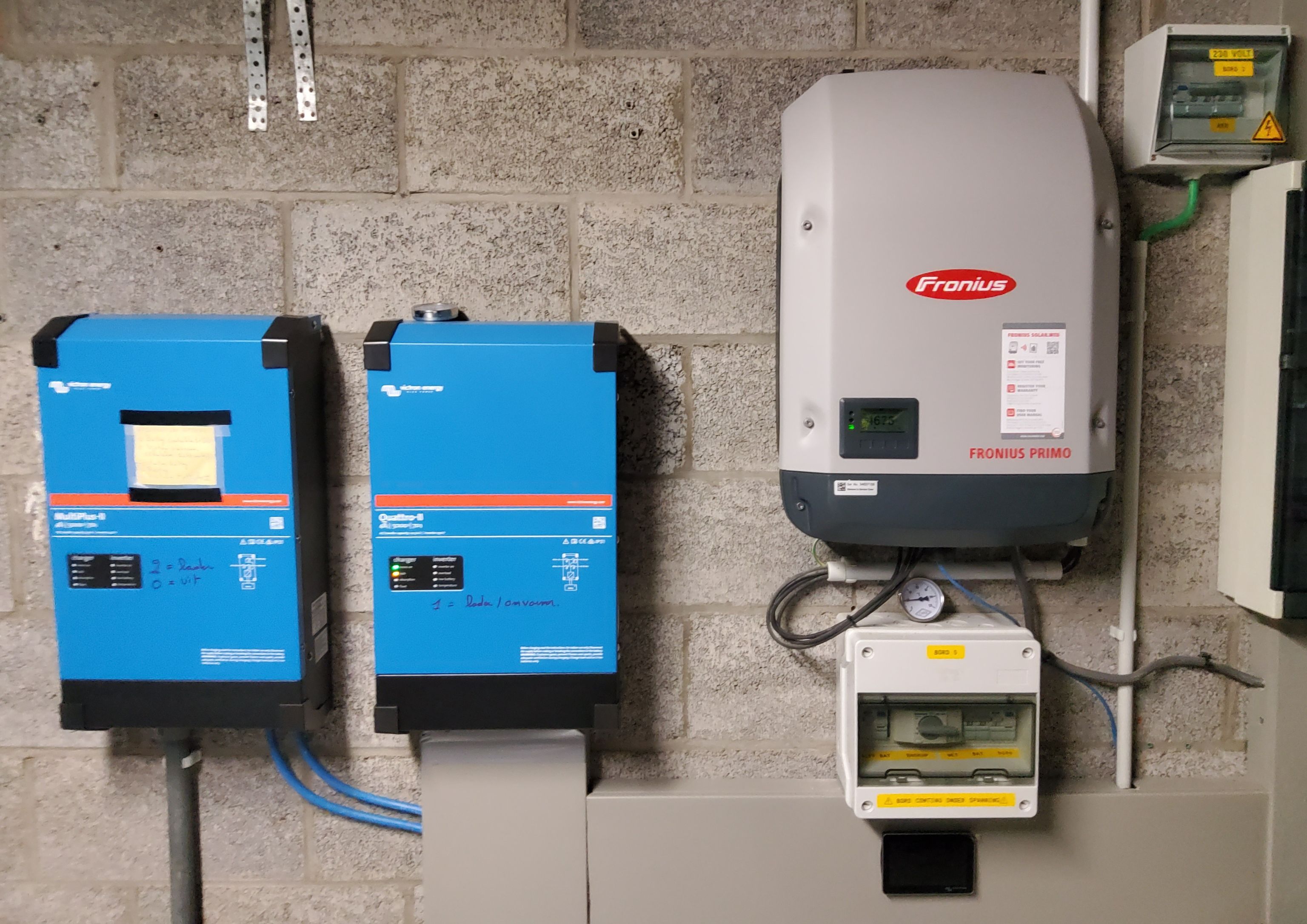 Electrical equipment mounted on a gray brick wall, including two blue devices and a gray inverter labeled "Fronius."