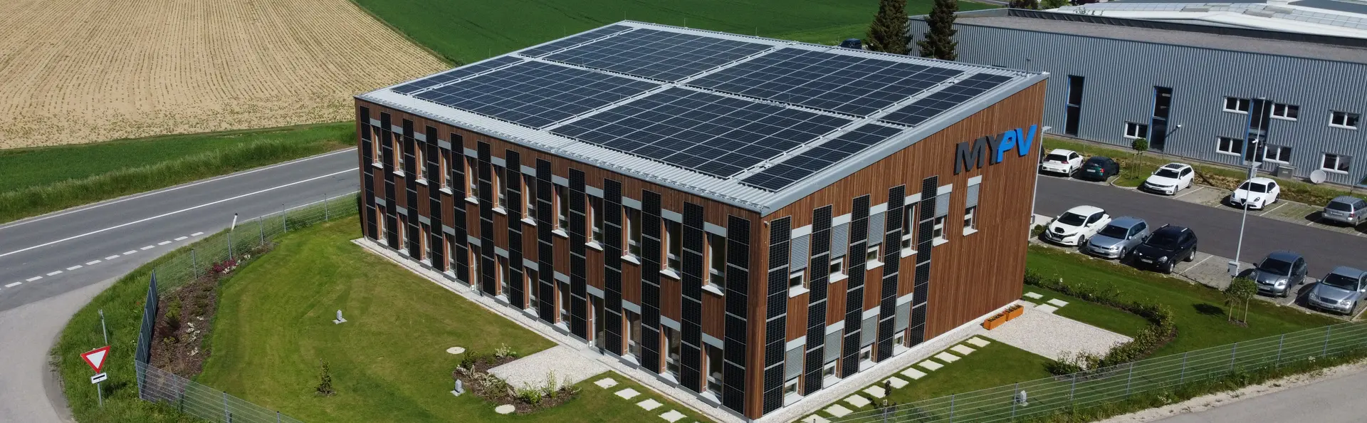 Office building of my-PV with PV modules on the roof and facade, surrounded by green space and parking lot.