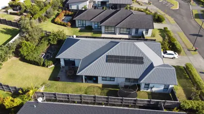 Aerial view of a modern house with a gray metal roof and solar panels, surrounded by a garden and driveway, located in a suburban neighborhood.