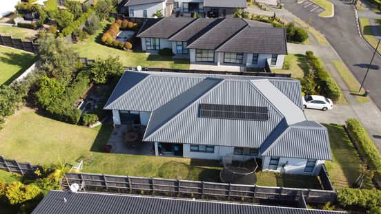 Aerial view of a modern house with a gray metal roof and solar panels, surrounded by a garden and driveway, located in a suburban neighborhood.