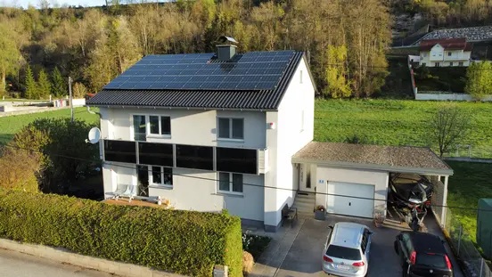 A house with solar panels on the roof, a garden enclosed by shrubs, and a garage with two cars parked outside.