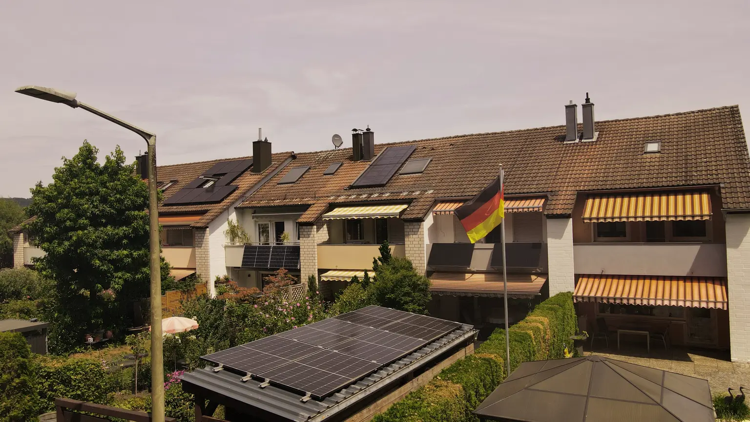 Row of houses with rooftop solar panels and a German flag, surrounded by lush greenery under a clear sky.