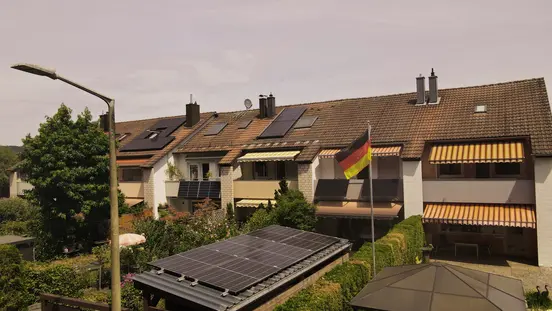 Rooftops with solar panels, German flag, tree, awnings, and a streetlamp on a sunny day.