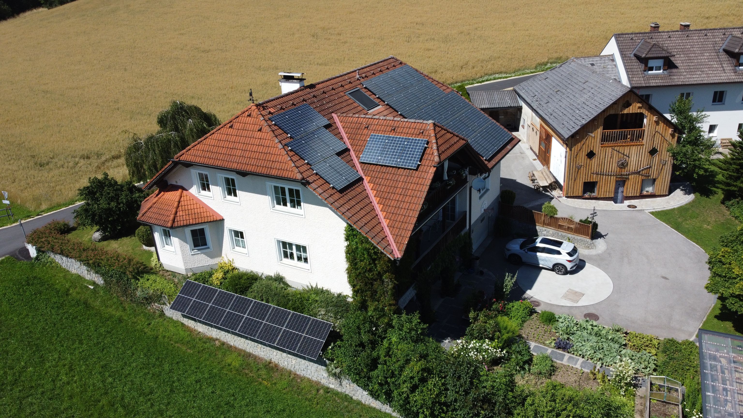 Aerial view of a house with solar panels on the roof, a garden and driveway, next to buildings, set against open fields.